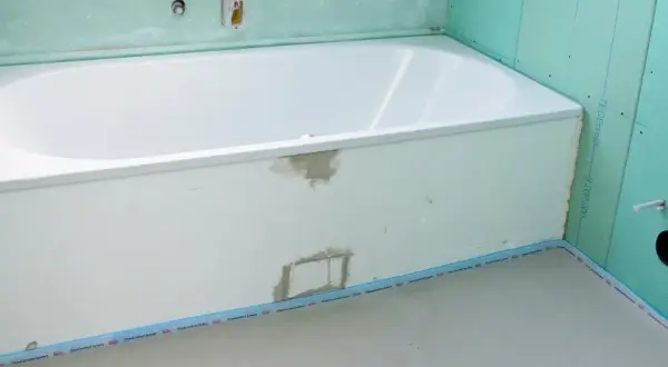 Einbau einer neuen, weißen Badewanne in einem Badezimmer im Rohbau. Grüne Gipskartonplatten und vorbereitete Rohre sind sichtbar.