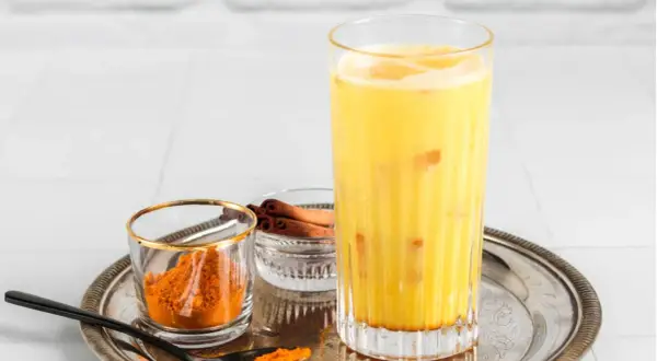 Ein Glas goldene Milch mit Kurkumapulver und Zimtstangen daneben.