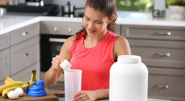Donna prepara un frullato con proteine prima o dopo allenamento, con banane e uova sul bancone.