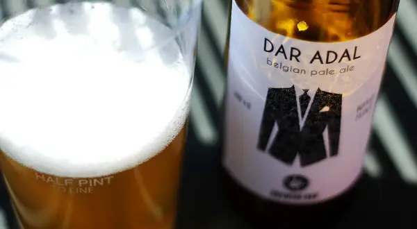 Bicchiere di birra dorata con schiuma bianca e bottiglia di Dar Adal belgian pale ale con etichetta a forma di giacca.