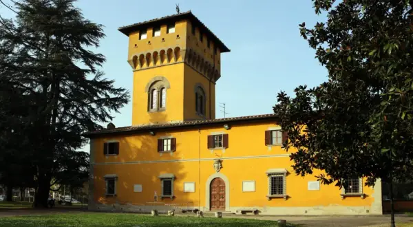 Edificio giallo con torre merlata a Borgo San Lorenzo, circondato da alberi verdi e un cielo azzurro.
