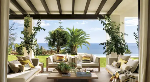 Cómodos porches de entrada principal con sofás de madera, cojines y mantas, con vistas al mar y palmeras.