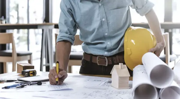 Ein Bauleiter, der als Generalunternehmer Baupläne prüft. Er hält einen gelben Helm und einen Stift.