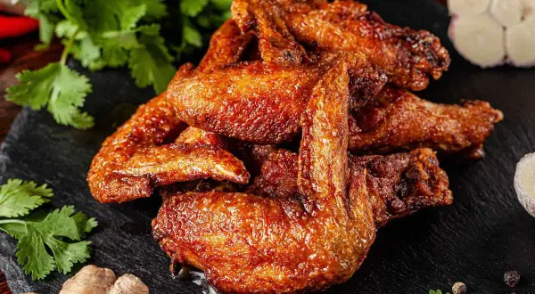 A pile of golden-brown, crispy alette di pollo, glistening with sauce, are served on a dark slate. Coriander, ginger, garlic, and peppercorns complete the appetizing presentation.