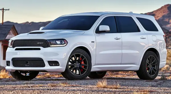 Biały Dodge Durango 3.6 SRT z czarnymi felgami i czerwonymi zaciskami hamulcowymi stoi na żwirowej drodze, w tle góry i opuszczony dom.