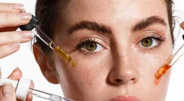 Hände tragen Pipetten mit Seren auf das Gesicht einer Frau auf. Diese Seren enthalten oft Peptide, die für die Hautpflege wichtig sind.