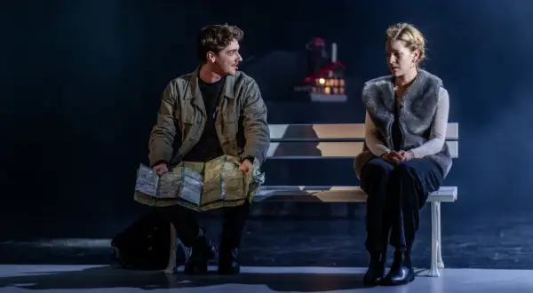 Para na ławce, mężczyzna z mapą, kobieta w futrzanej kamizelce. Scena z dramatu "Jelegnia Góra" pełna napięcia.