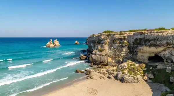 Scogliere calcaree e mare turchese, un paradiso tra le località balneari puglia. Faraglioni emergono dall'acqua cristallina, mentre una grotta si apre nella roccia.