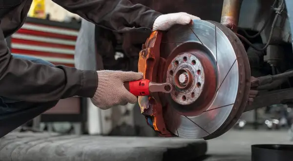 Mechanik w rękawiczkach dokonuje wymiany klocków hamulcowych, używając klucza do odkręcenia śruby zacisku.