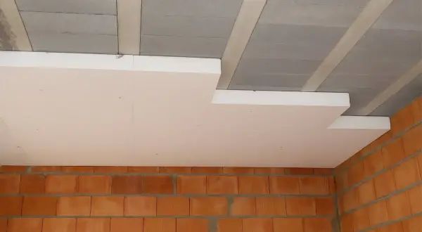 Białe płyty styropapa montowane pod betonowym stropem, nad ceglanymi ścianami. Izolacja termiczna w trakcie budowy.