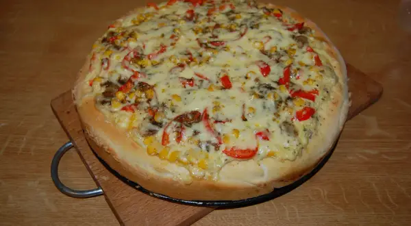 Pizza z kukurydzą, papryką i grzybami, polana obficie białym sosem do pizzy, na drewnianym blacie.