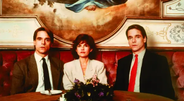 Jeremy Irons w filmach, tutaj z dwiema innymi osobami, siedzi przy stole w eleganckiej restauracji.