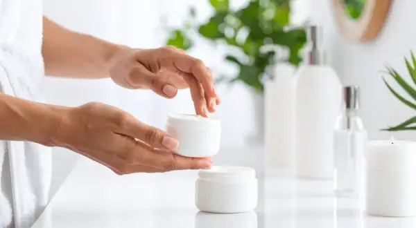 Dłonie nakładają krem z PEG-40 hydrogenated castor oil na twarz. W tle kosmetyki i rośliny.
