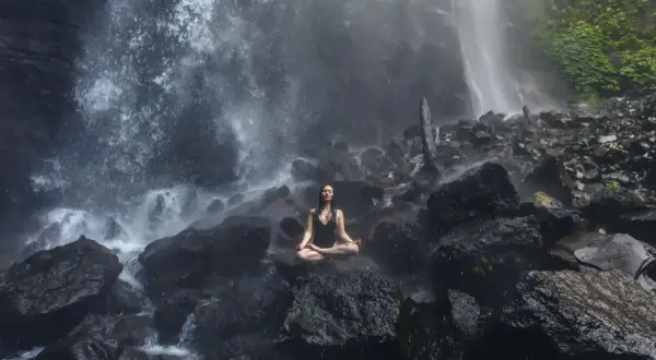 Mujer meditando en posición de loto sobre rocas, rodeada por la bruma de una cascada. Un momento de sanación pránica en la naturaleza.