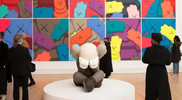 A KAWS sculpture sits before a vibrant mural, inspiring KAWS painting ideas. Visitors admire the art.