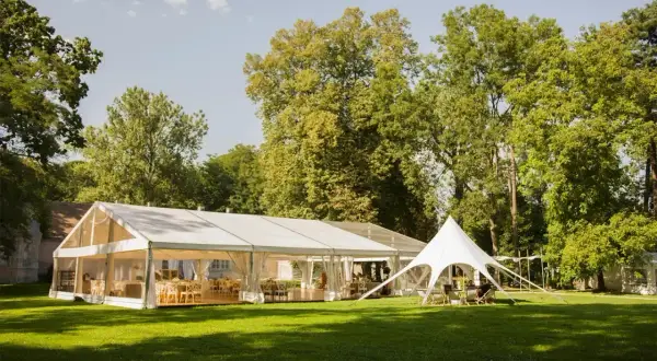 Eleganckie wesele w namiocie na trawie, wśród drzew. Wnętrze namiotu udekorowane stołami i krzesłami, gotowe na przyjęcie gości.