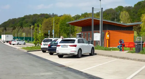 Stationnement sur une aire d'autoroute avec plusieurs voitures garées. Un homme est assis sur un banc rouge devant un bâtiment orange.