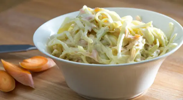 En skål med krämig, godaste coleslaw med strimlad vitkål och morötter. Bredvid ligger några morotsskivor.