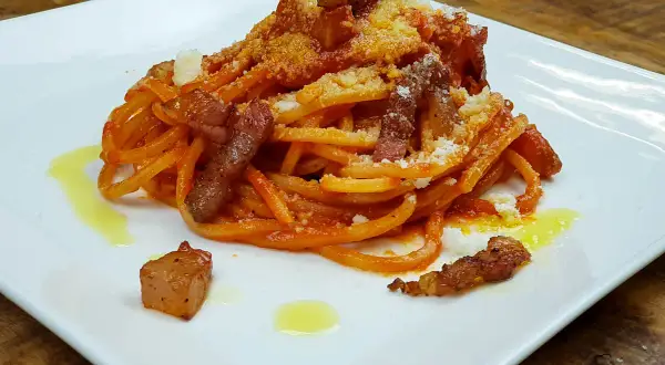 Bucatini con salsa amatriciana, cubos de guanciale crujiente y queso rallado, servido en plato blanco.