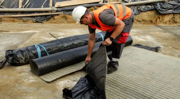 Budowlaniec rozkłada warstwę **betonitu** na placu budowy. Obok leżą zrolowane materiały izolacyjne.