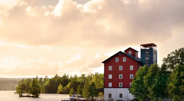Skålleruds gård vid vattnet, med en röd byggnad och ett torn, omgiven av träd under en molnig himmel.