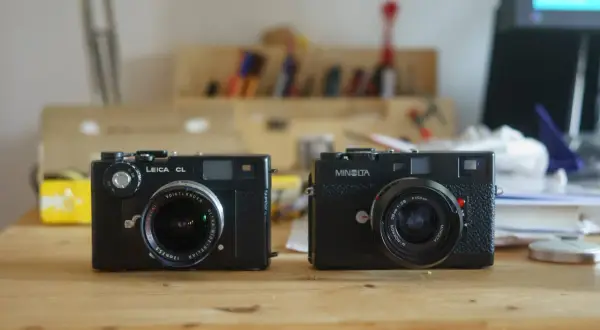 Two classic rangefinder cameras, a Leica CL and a Minolta, sit side-by-side on a wooden desk, ready for their next adventure.