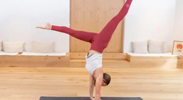 Mujer en postura de **Adho Mukha Vrksasana** (pino) con piernas abiertas en V, en un estudio de yoga.
