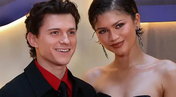 Tom Holland y Zendaya sonríen juntos en un evento. Él lleva una camisa roja y corbata negra. Ella luce un vestido negro y pendientes largos.