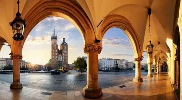 Krakau Geheimtipps: Blick durch Arkaden auf den Marktplatz mit Marienkirche im Sonnenaufgang. Ein malerischer Ort für Entdeckungen.
