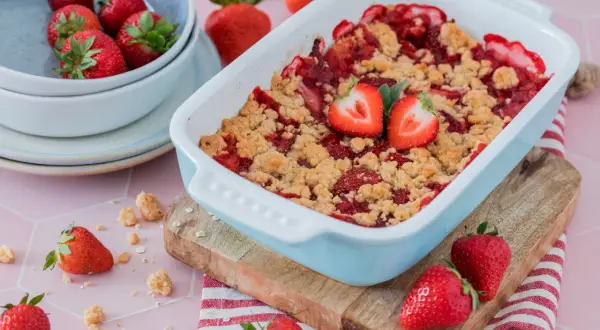 Ein frisch gebackener Erdbeer Crumble in einer hellblauen Auflaufform, garniert mit Erdbeerhälften. Daneben eine Schüssel mit Erdbeeren und verstreute Streusel.