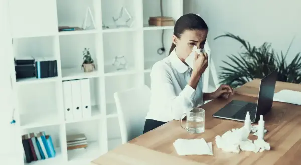 Kobieta z chusteczką przy nosie siedzi przy biurku z laptopem. Na stole leżą leki i chusteczki. To chyba jej ostatni dzień pracy, l4 na wypowiedzeniu.