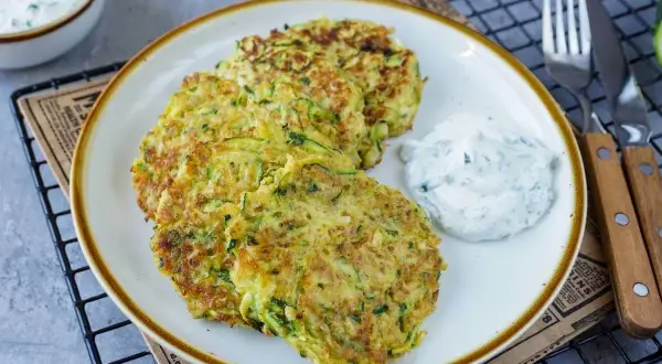 Drei goldbraune Zucchini-Pfannkuchen liegen auf einem Teller, daneben ein Klecks Kräuterquark. Ein leckeres Gericht!