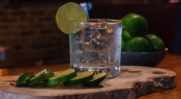Erfrischender Gin Tonic Water mit Eis und Limettenscheiben auf einem Holzbrett. Mehrere Limetten liegen in einer Schale daneben.