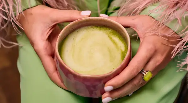 Dłonie trzymają kubek z zieloną matchą, zastanawiając się, czy matcha odchudza. Ubrana w pistacjowy strój z różowymi piórami.