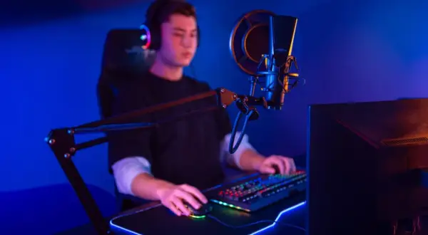 Ein junger Mann mit Kopfhörern sitzt vor einem beleuchteten Mikrofon und einer Tastatur. Er ist ein Twitch Streamer, der seine Spiele oder Inhalte live teilt.