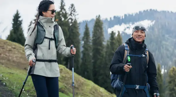 Para w odpowiednim ubraniu w góry, z kijkami trekkingowymi, cieszy się wędrówką pośród zielonych wzgórz i ośnieżonych szczytów.