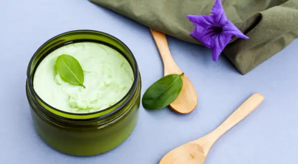 Un pot de gommage cuir chevelu maison, vert pâle, avec des cuillères en bois et une fleur violette.