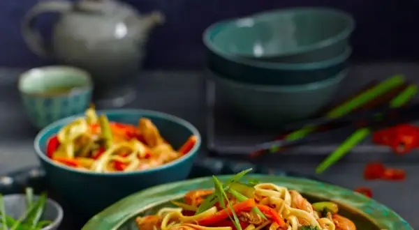 Makaron Chow Mein z kurczakiem i warzywami w misce, na tle czajniczka i pałeczek.
