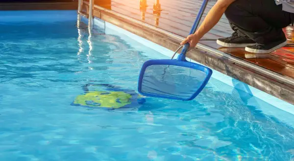 Person mit Kescher im Wasser, um den Pool sauber machen. Ein gelbes Spielzeug treibt im klaren Wasser.
