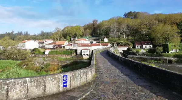 Antiguo puente de piedra con señal de peregrinación. Al fondo, un pueblo con casas de tejados rojos y vegetación exuberante. ¡Ponte Maceira que ver!