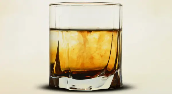 Szklanka z bursztynowym płynem, w którym rozpuszcza się ciemniejsza substancja, przypominająca proces, jak zrobic whiskey.