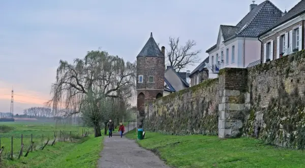 Ein Paar spaziert auf einem Weg entlang der alten Stadtmauer von Zons am Rhein. Ein Weidenbaum und ein Wehrturm prägen die Szenerie.