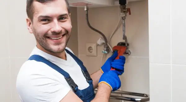 Uśmiechnięty hydraulik w niebieskim kombinezonie i rękawiczkach dokręca kluczem bojler. Zastanawiasz się, czy wyłączać bojler na wyjazd? On wie!