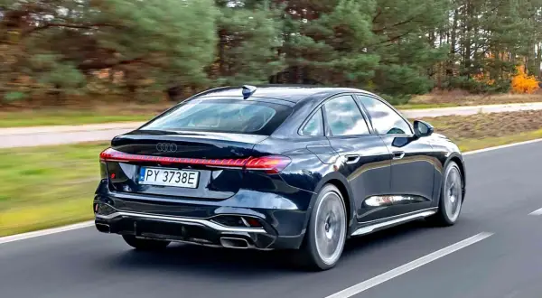Ciemny Audi A4 B10 w wersji sedan pędzi po drodze, otoczony jesiennym lasem.