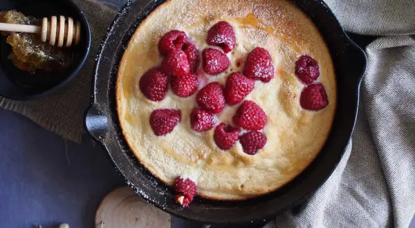 Puszysty omlet z malinami, posypany cukrem pudrem, podany w żeliwnej patelni. Obok miseczka z miodem i plastrem pszczelim.