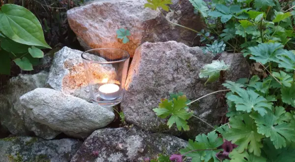 Ein Teelicht in einem Glas leuchtet zwischen Steinen und grünen Pflanzen. Ein Symbol für Hoffnung und einen bibelvers mut.