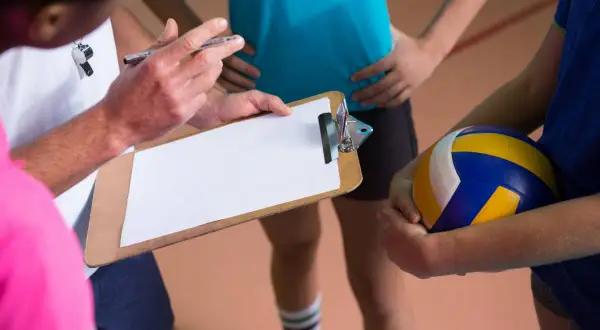 Trener omawia z drużyną błąd ustawienia w siatkówce, trzymając tablicę z notatkami.
