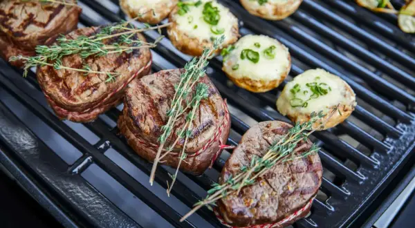 Saftige Rindersteaks mit Kräutern auf dem Holzkohlegrill. Daneben liegen gefüllte Champignons.