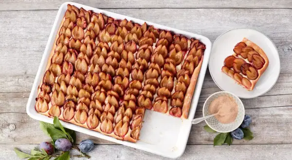 Ein köstlicher Zwetschgenkuchen vom Blech, mit saftigen Pflaumen belegt und goldbraun gebacken. Daneben ein Stück Kuchen auf einem Teller.