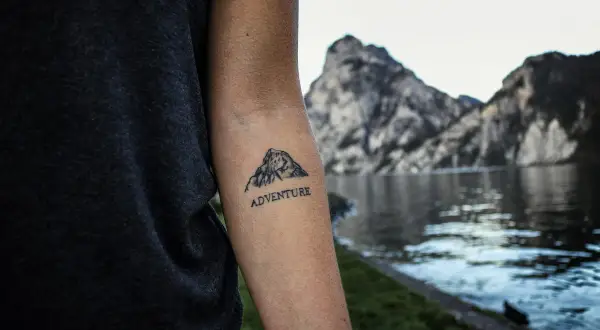 Tatuaż góry z napisem "ADVENTURE" na ramieniu, na tle jeziora i skalistych szczytów.